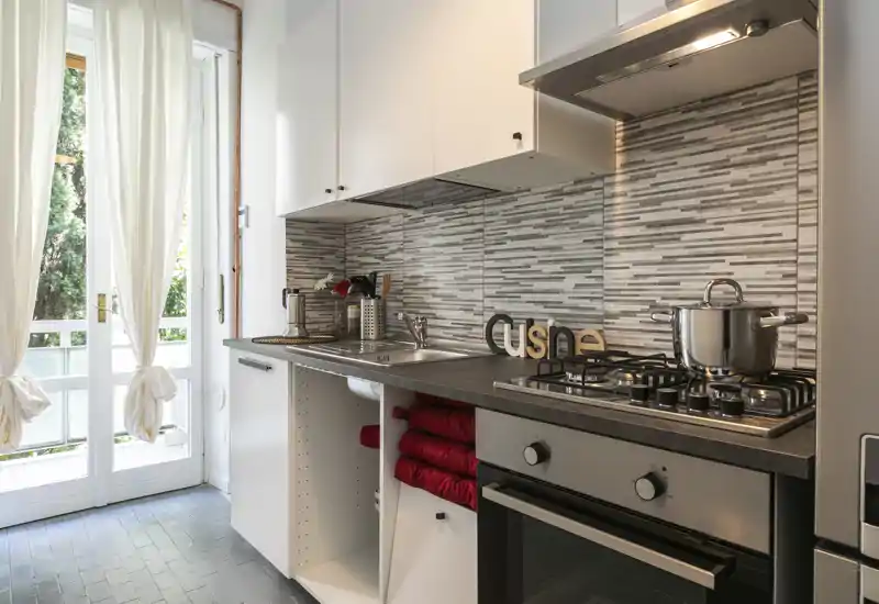 Modern compact kitchen with gas stove, oven and stainless sink, bright natural light from French doors and a decorative tiled backsplash; neatly folded red towels and small accents create a welcoming feeling.