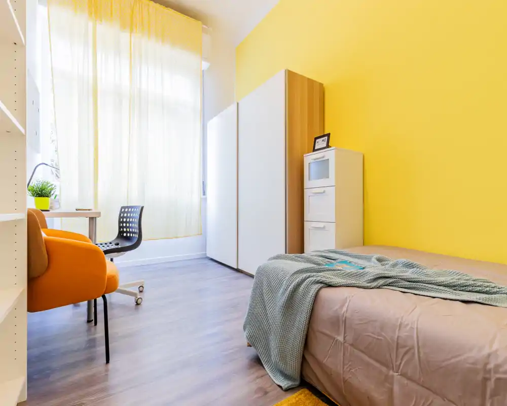 Camera da letto singola moderna e luminosa con letto ben rifatto, plaid, armadio e piccolo scrittoio vicino a una grande finestra che illumina la stanza.