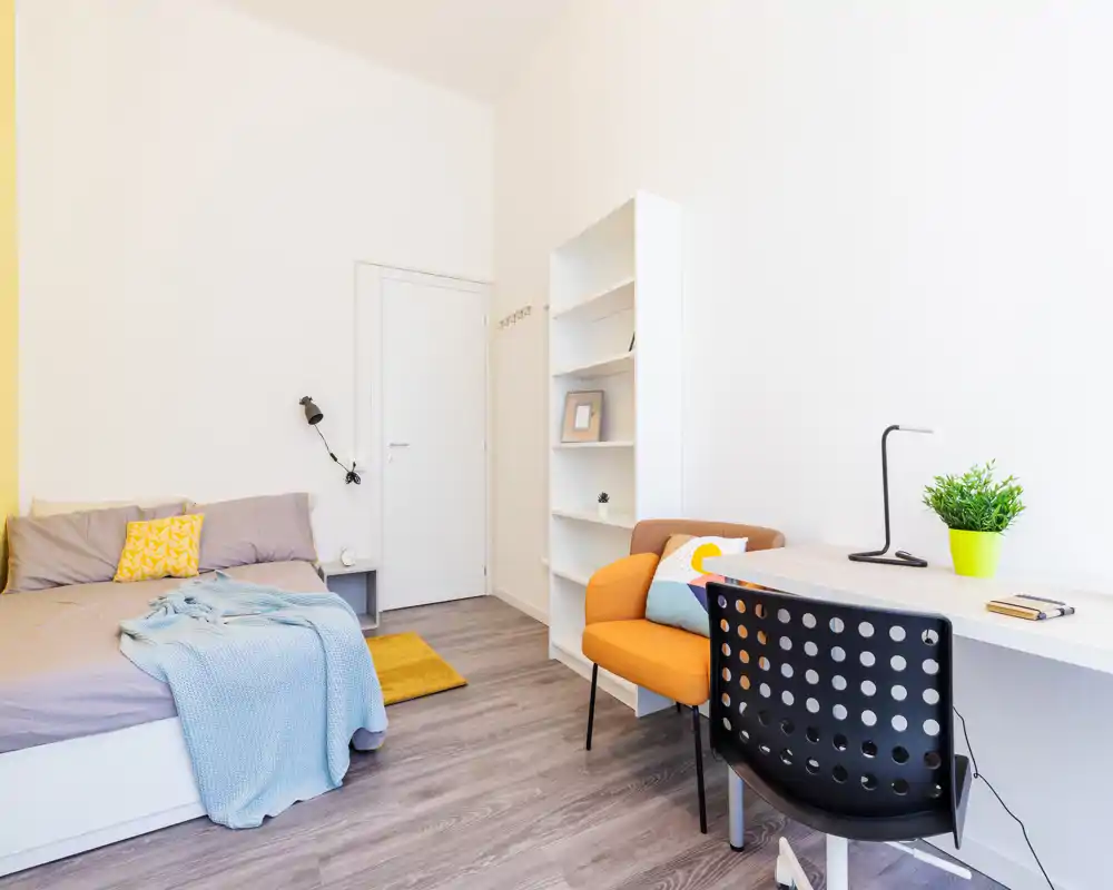 Camera da letto luminosa e moderna con letto matrimoniale ben rifatto, tessili accoglienti e un angolo studio con scrivania e sedia — perfetta per un soggiorno confortevole.