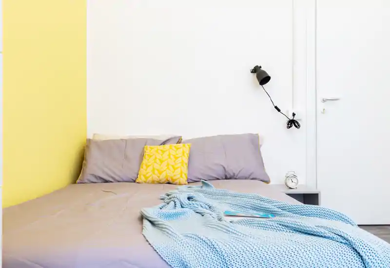 Camera da letto luminosa e moderna con letto rifinito, cuscini decorativi e un morbido plaid lavorato. Palette bianca e gialla e una lampada minimale creano un'atmosfera fresca e accogliente.