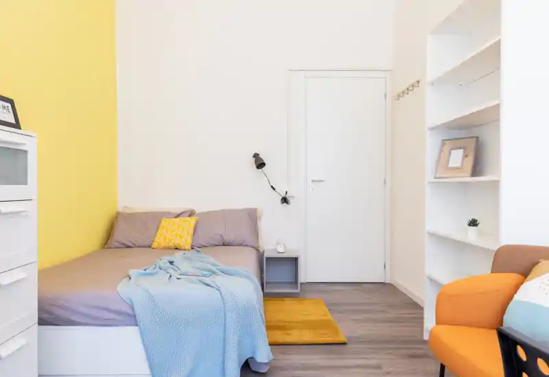 Camera da letto luminosa e accogliente con letto ben rifatto, plaid azzurro e cuscino giallo; scaffalatura moderna e poltrona calda completano l'ambiente.