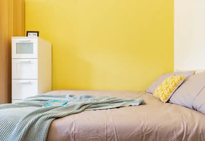 Camera da letto luminosa e moderna con letto ben rifatto, cuscini decorativi e una coperta lavorata a maglia davanti a una parete accentata gialla.