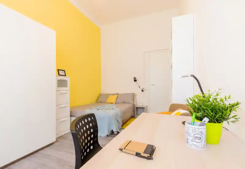 Camera da letto luminosa e moderna con parete gialla, letto matrimoniale con biancheria neutra e cuscini decorativi, e una scrivania in primo piano — ideale per studenti o professionisti.