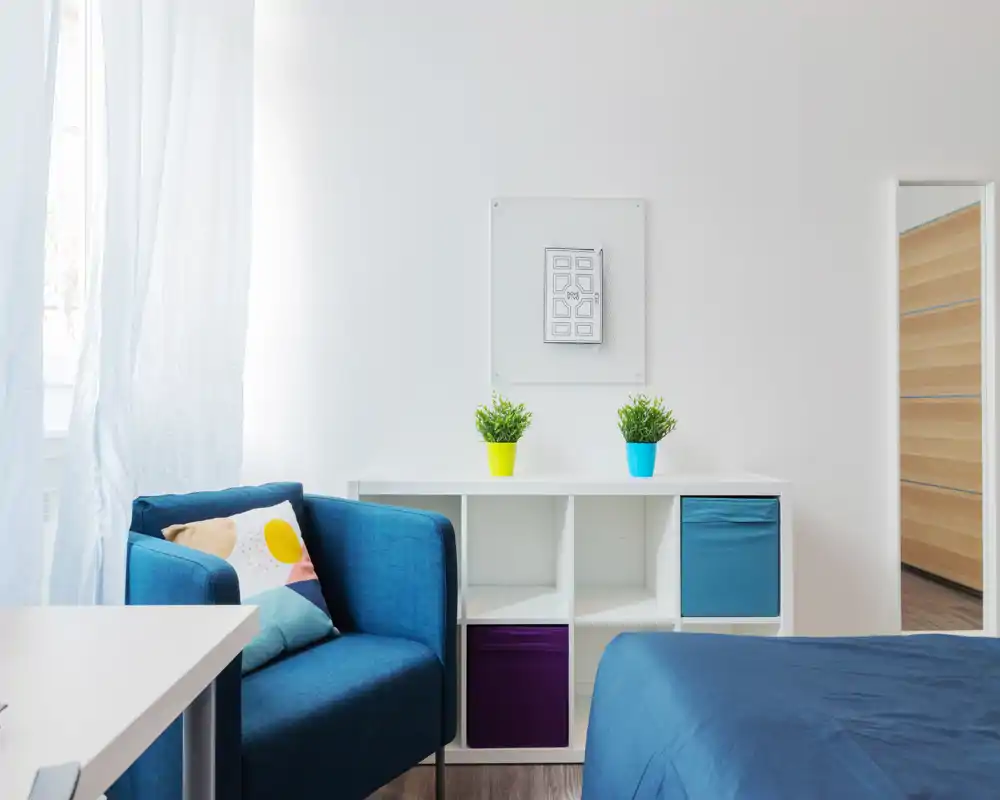 Bright, modern bedroom corner with a blue armchair, compact shelving and a neatly made bed. Natural light, colorful cushions and small plants create a fresh, welcoming feel.