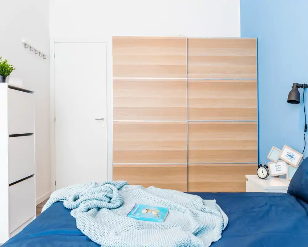 Bright, modern bedroom with a neatly made bed, soft knit throw and light wood wardrobe creating a calm, airy atmosphere.