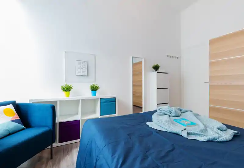Dormitorio moderno y luminoso con cama azul bien hecha, manta acogedora y plantas decorativas; mobiliario minimalista y un pequeño sofá azul crean un espacio acogedor.