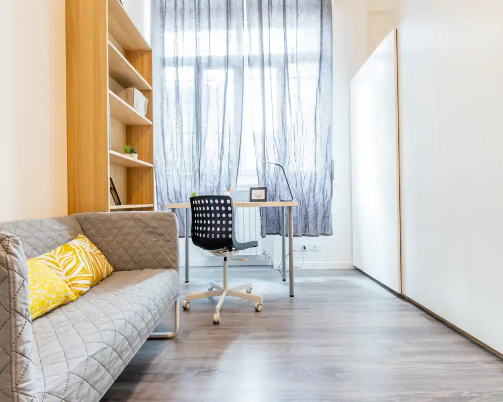 Bright, modern living area with a compact sofa, decorative shelving and a small desk by a large window—ideal for relaxing or working in a cozy, well-lit space.