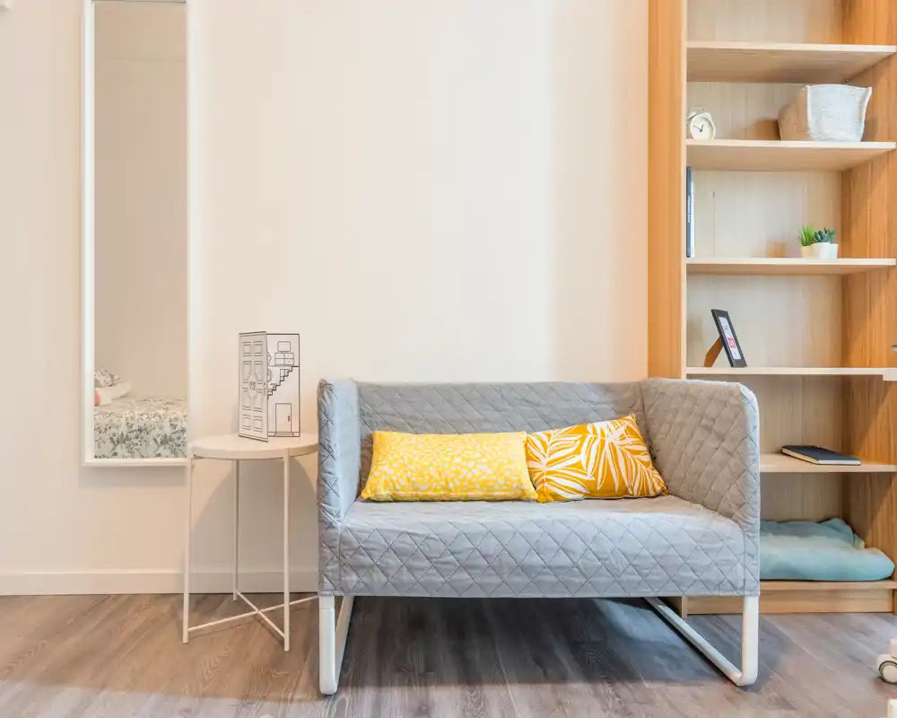 Bright, minimalist living area with a quilted two-seater sofa, decorative yellow cushions and open wooden shelving — a cozy seating nook ideal for a compact apartment.