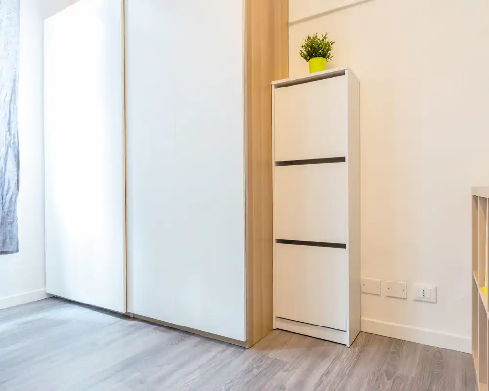 Image shows a corner of a room with a wardrobe/closet, a slim storage cabinet with a small plant on top, and bare floor; no clear bed, seating, kitchen or bathroom fixtures are visible.
