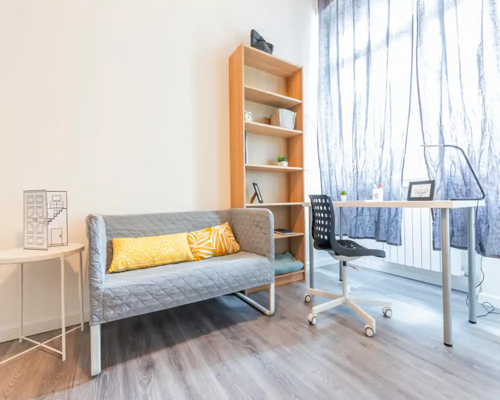 Bright, modern living area with a compact grey sofa, decorative cushions and a workspace by a large window letting in soft natural light.