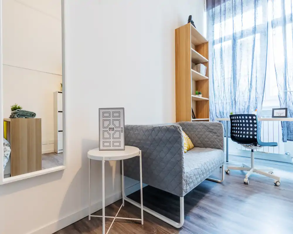 Modern, bright studio corner with a compact quilted sofa, study desk by a large window and a slim shelving unit — a cozy, well-lit living/work nook ideal for a city rental.