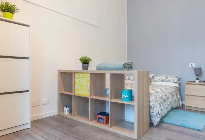 Bright, modern bedroom corner with a double bed dressed in patterned linens, a small wooden bedside table and an open shelving unit that separates the sleeping area from the rest of the room.