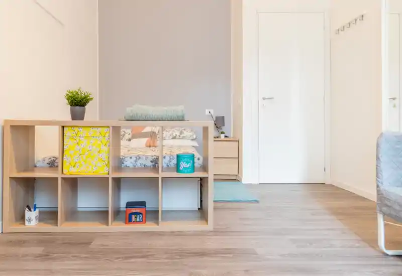 Bright, modern bedroom partially framed behind a low shelving unit; visible pillows, patterned bedding and a bedside table create a tidy, welcoming sleeping area.