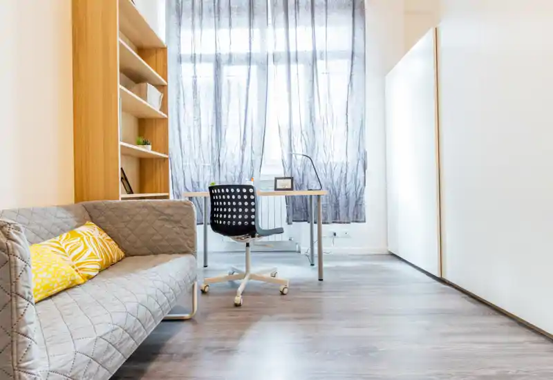 Bright, modern living area with a compact sofa, decorative shelving and a small desk by a large window—ideal for relaxing or working in a cozy, well-lit space.