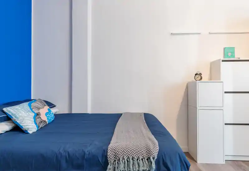 Dormitorio moderno y luminoso con cama bien hecha, cojines decorativos y manta texturizada. Líneas limpias, pared azul como acento y mueble blanco minimalista crean un ambiente ordenado y atractivo.
