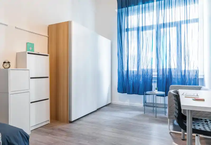 Rincón de dormitorio luminoso y moderno con armario compacto, pequeño escritorio y cortina azul translúcida que deja pasar la luz natural; estilo minimalista y suelo laminado limpio.
