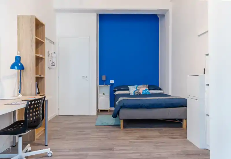 Dormitorio moderno y luminoso con pared de acento azul intenso, cama doble cómoda, mesita de noche y escritorio. Líneas limpias y textiles coordinados hacen la habitación acogedora y funcional para descansar y trabajar.