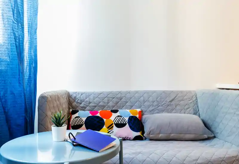 Área de estar luminosa y moderna con sofá gris acolchado, cojín decorativo colorido, mesita redonda y una planta — un rincón acogedor para relajarse.