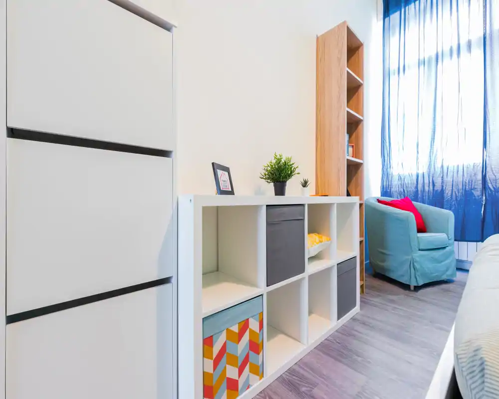 Rincón de dormitorio luminoso y moderno con estanterías abiertas, un butaca tapizado confortable junto a la ventana y luz natural suave; almacenamiento ordenado y detalles decorativos que crean un ambiente acogedor.