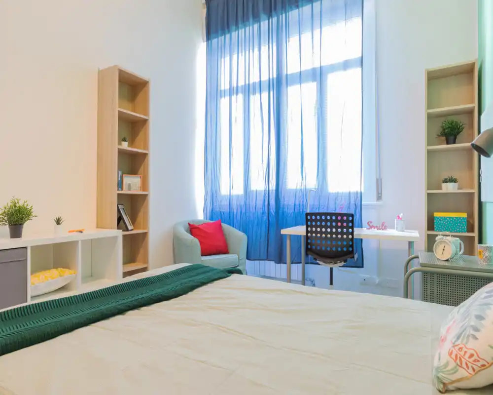 Dormitorio luminoso y decorado, con cama grande en primer plano, escritorio junto a la ventana y detalles acogedores como sillón, cojines y plantas.