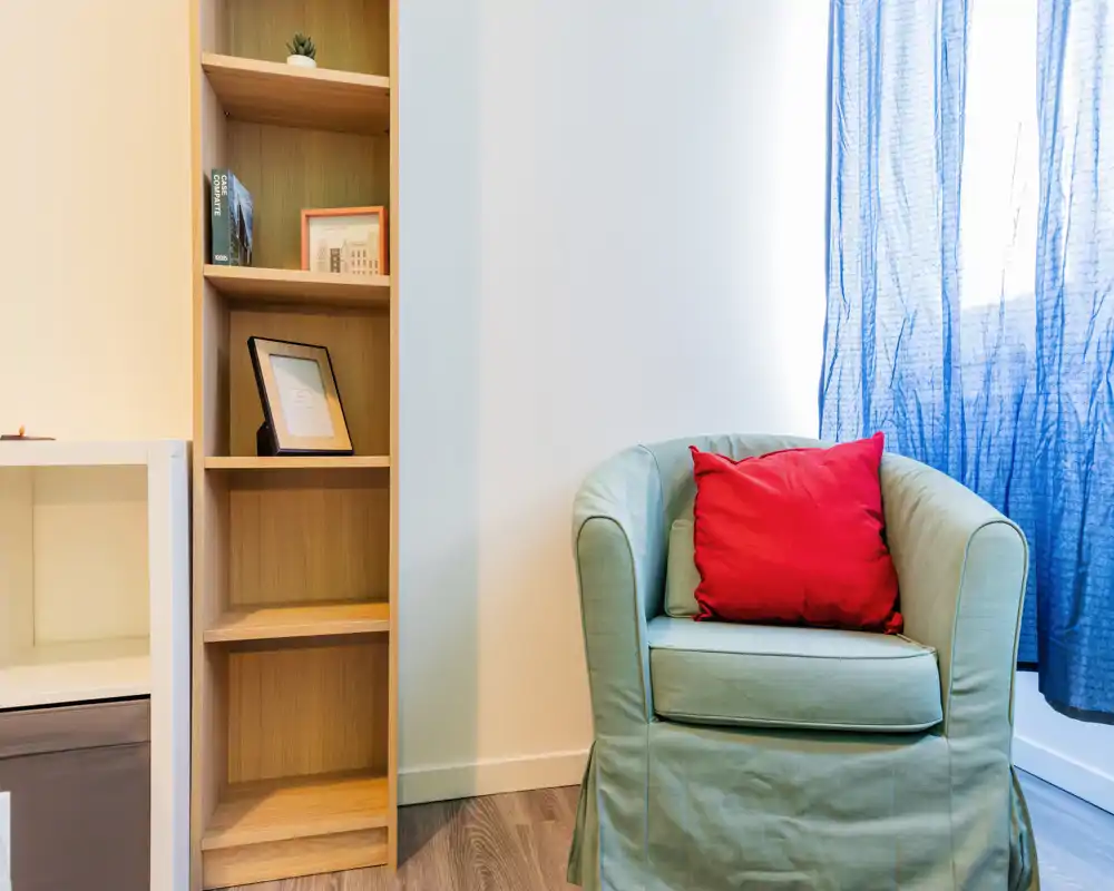 Rincón de salón acogedor con un sillón tapizado verde claro y un cojín rojo brillante junto a una estantería de madera estrecha y una ventana con cortina. La escena está bien iluminada y pensada para un rincón de lectura.