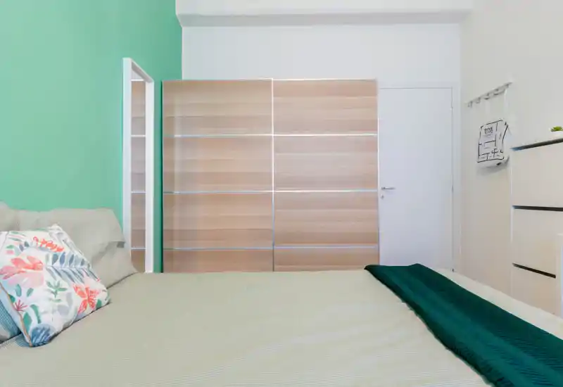 Dormitorio luminoso y ordenado con una cama grande en primer plano, pared verde, armario corredero de madera y cojines decorativos que aportan una sensación acogedora.