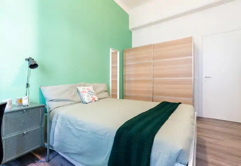 Dormitorio luminoso y bien dispuesto con cama doble, cojín decorativo y pared verde; armario amplio y mesita visibles, ideal para anuncios de alquiler.