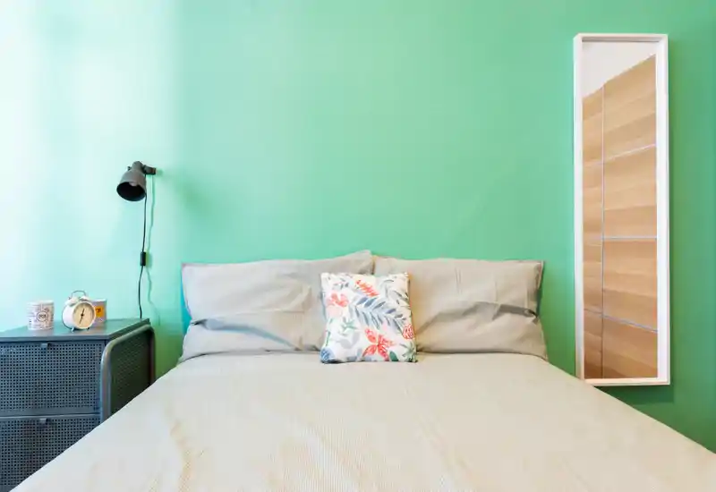 Dormitorio luminoso y ordenado con cama completa centrada frente a una pared verde menta, cojín floral y mesita con lámpara y reloj despertador; perfecto para fotos de alquiler que muestren una zona de descanso acogedora.