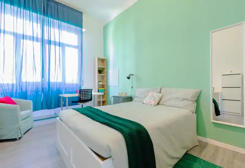 Dormitorio luminoso y moderno con cama grande, pared verde menta, escritorio junto a la ventana y un sillón acogedor, perfecto para descansar o trabajar.