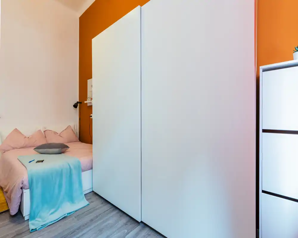 Camera da letto luminosa e accogliente con letto matrimoniale con biancheria rosa, cuscini e un plaid azzurro; grande armadio bianco in primo piano per ampio spazio di stoccaggio.