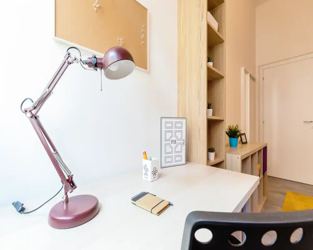 Angolo di soggiorno moderno e luminoso con scrivania ordinata, lampada regolabile e scaffalature. Toni neutri del legno, piccole piante e organizzazione creano uno spazio accogliente e funzionale, ideale per smart working o lettura.