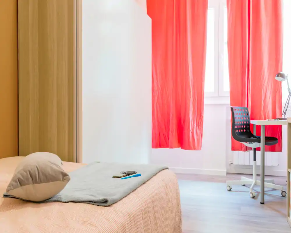 Camera da letto luminosa e semplice con un letto confortevole in primo piano, tessili morbidi e una scrivania vicino alla finestra con tende rosse che filtrano la luce naturale.