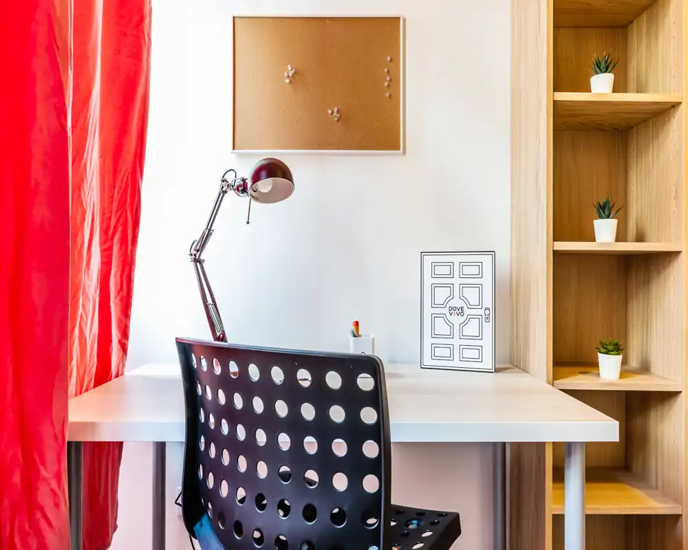 Angolo studio luminoso in zona giorno: scrivania bianca con lampada da lavoro, sedia nera traforata, bacheca e scaffalatura in legno con piccole piante — spazio ordinato e funzionale vicino a una tenda rossa.