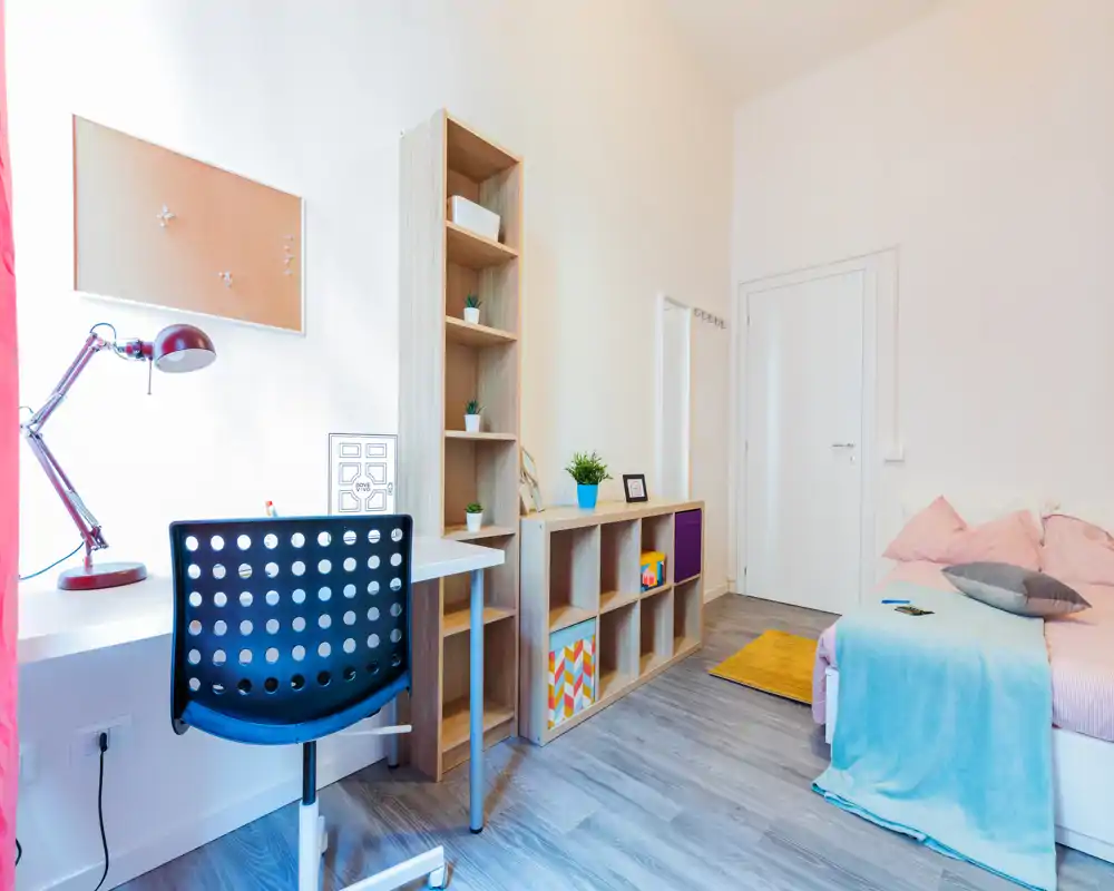 Camera da letto compatta e luminosa con letto singolo in lenzuola pastello, scrivania con lampada e sedia ergonomica e scaffalatura aperta per riporre oggetti — ideale per uno studente o una singola persona.