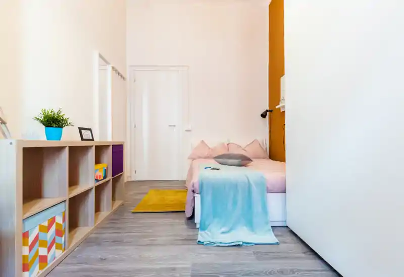 Camera da letto singola luminosa e curata, con letto in tonalità pastello, coperta morbida e cuscini, mobile basso contenitivo e accenti di colore caldi — perfetta per un affitto accogliente.