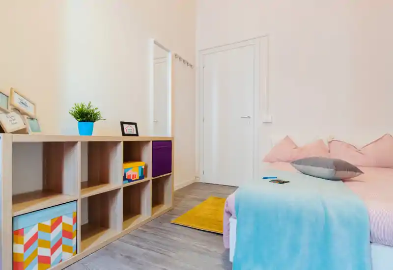 Camera da letto luminosa e ordinata con letto in tonalità pastello, cuscini, plaid azzurro e una scaffalatura ben organizzata: ambiente moderno e accogliente ideale per affitti brevi.