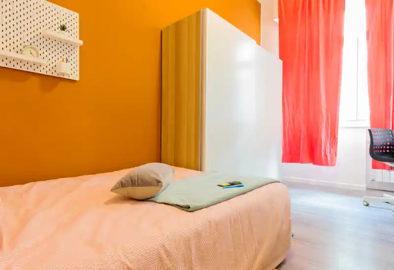Camera singola luminosa e ordinata con letto confortevole, parete accentata arancione e armadio vicino alla finestra che lascia entrare luce naturale.