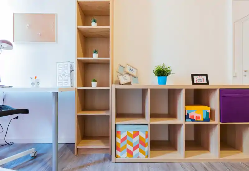 Zona de estar luminosa y moderna con estanterías modulares de madera, cajas decorativas y un pequeño escritorio: un espacio limpio y bien iluminado ideal para relajarse o trabajar.