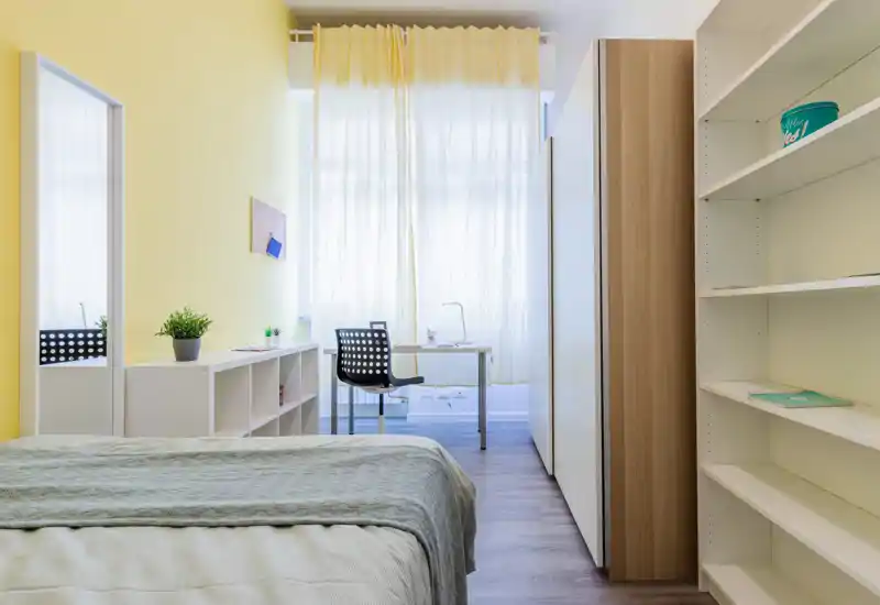 Dormitorio moderno y luminoso con la cama ordenada en primer plano, un escritorio junto a una amplia ventana con cortina, estanterías y una pared de acento amarilla.