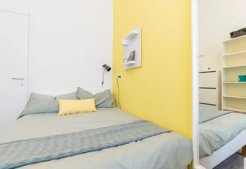Dormitorio moderno y luminoso con cama doble, ropa de cama verde suave y una pared amarilla que aporta un ambiente acogedor.