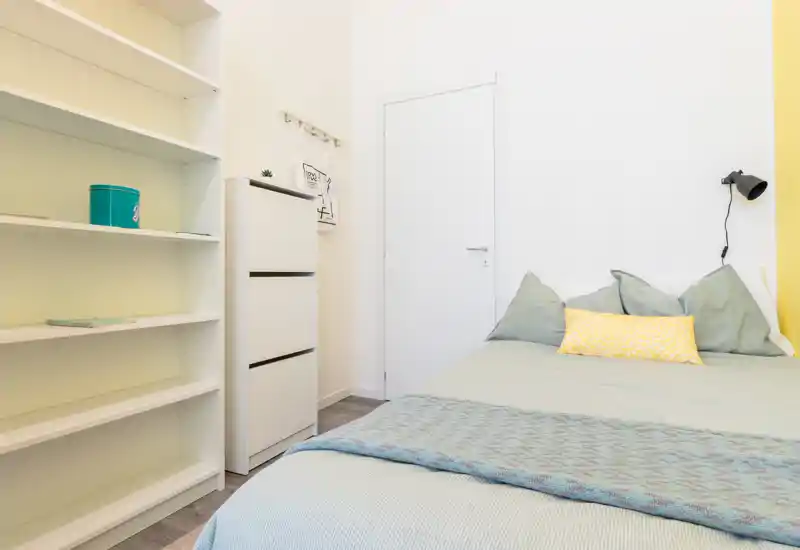 Dormitorio luminoso y minimalista con una cama doble bien hecha, cojines decorativos y una manta; estanterías y cómoda ofrecen almacenamiento y un aspecto moderno.