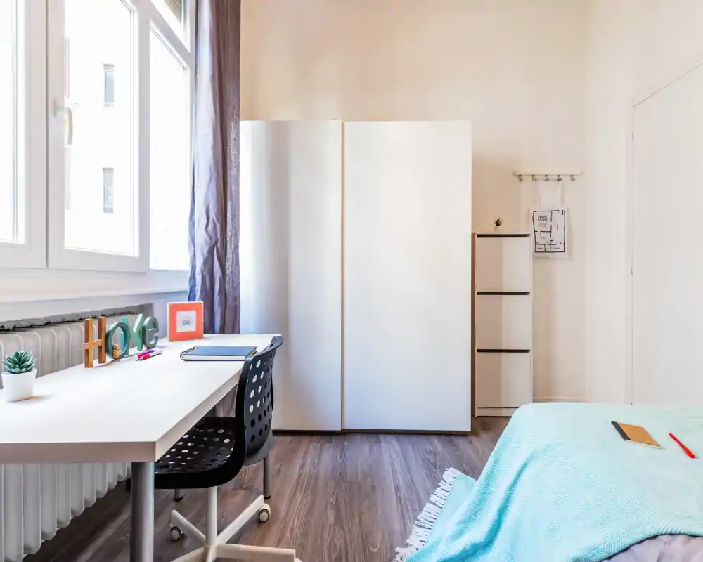 Camera da letto luminosa e minimal con scrivania vicino alla finestra, armadio bianco e letto parzialmente visibile con coperta azzurro chiaro.