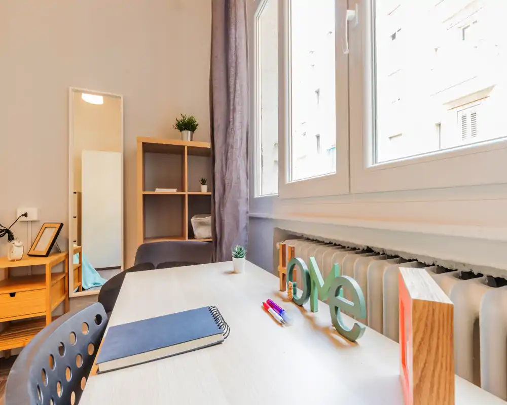Zona giorno luminosa e ordinata con una scrivania vicino a una grande finestra, elementi decorativi e mobili in legno che creano un’atmosfera accogliente.