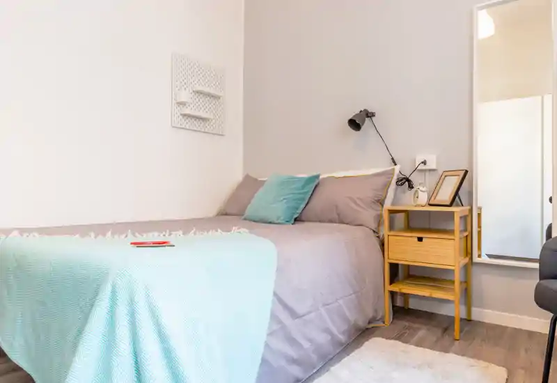 Camera da letto luminosa e ben curata con un comodo letto matrimoniale, copriletto pastello e comodino con lampada — uno spazio accogliente ideale per rilassarsi.