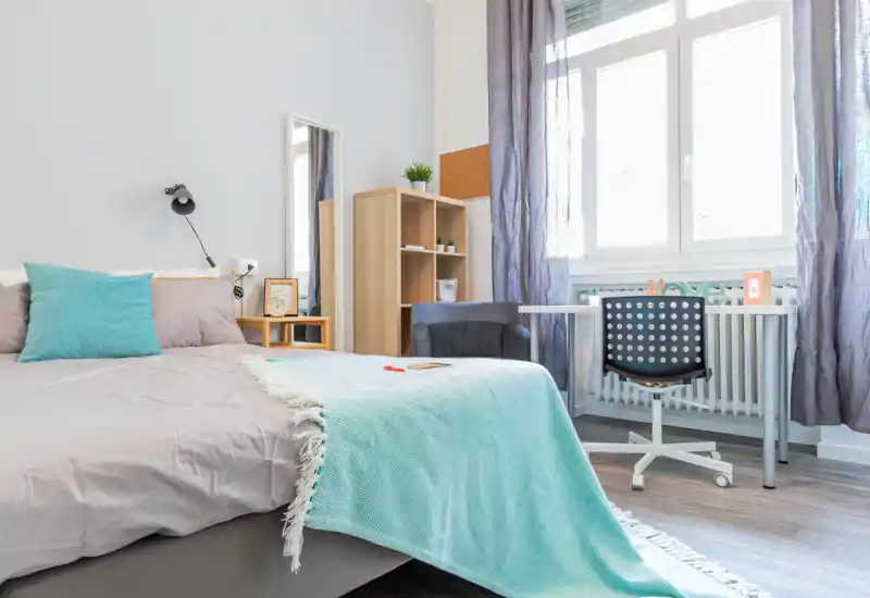 Camera da letto luminosa e ben arredata con grande letto con biancheria morbida, lampada da comodino e scrivania vicino alla finestra — perfetta per riposo e lavoro.