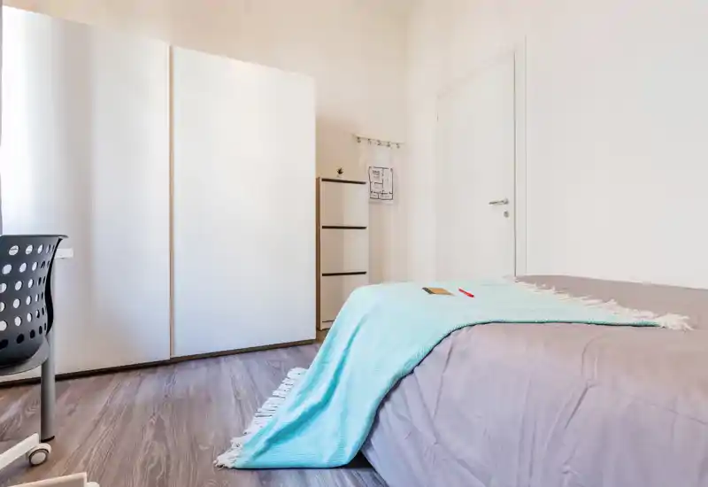 Camera da letto luminosa e minimal con letto ben rifatto con un plaid azzurro, grande armadio bianco e una sedia da scrivania — spazio ordinato e ben illuminato ideale per una persona.