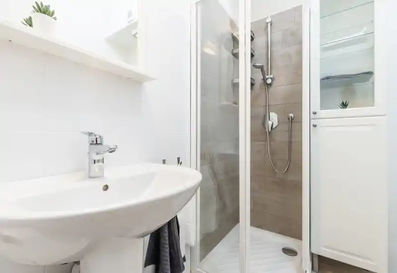 Modern compact bathroom with a pedestal sink, chrome faucet and a corner shower with glass doors and wood-effect tiles; white storage cabinet and bright, clean finishes.