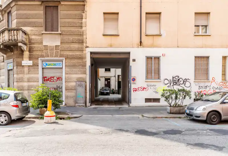 Street-facing exterior with an open passageway leading into a courtyard; parked cars, graffiti, and planters indicate an urban residential frontage.