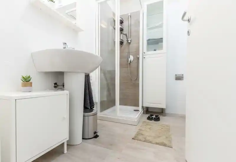 Bright, modern bathroom with a corner glass shower, pedestal sink, storage cabinet and light wood-effect flooring — clean and minimal.