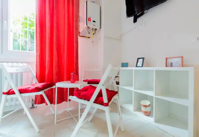 Small, bright living area with white folding chairs, a low round table and vibrant red cushions and curtains creating a cozy seating corner.